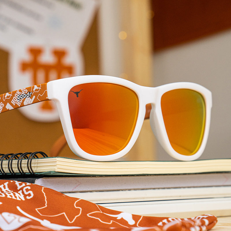 White-framed sunglasses with orange reflective lenses, featuring Texas Longhorns branding on the arm, resting on a stack of notebooks and surrounded by Texas-themed decor.