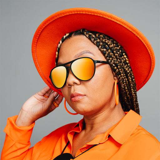 A woman poses fiercely in an orange hat and shirt wearing black aviator sunglasses with amber lenses.