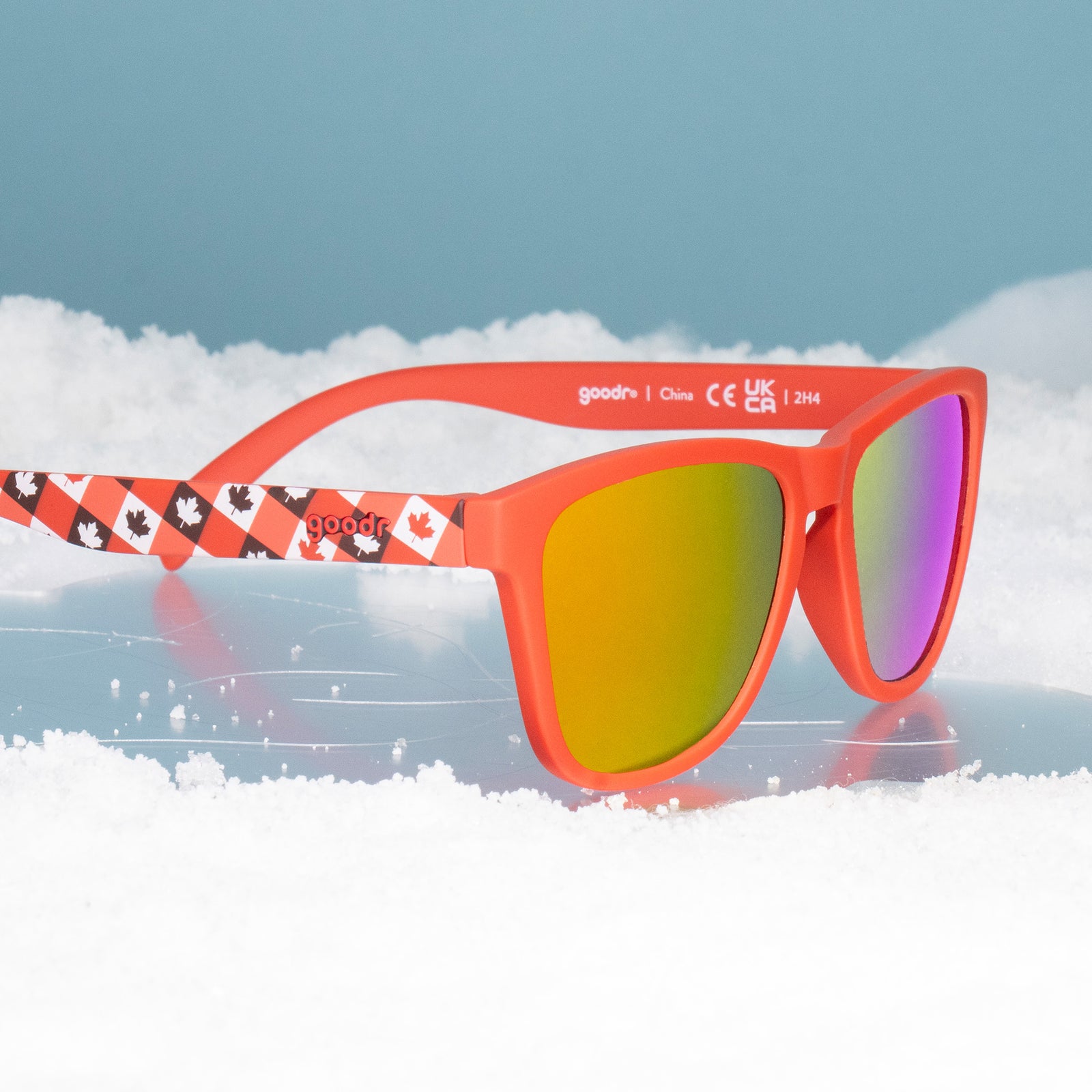 Three-quarter angle view of red maple-leaf patterned Canada sunglasses sitting on a patch of ice surrounded by snow.