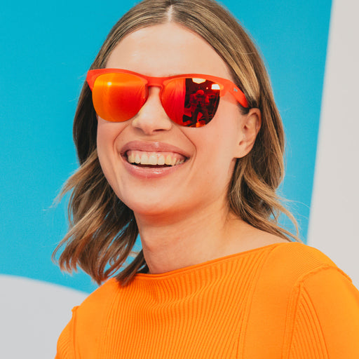 A stylish woman wearing vibrant red-orange half-rim sunglasses with red-orange reflective lenses looks off.