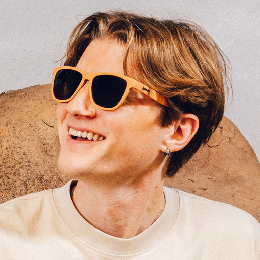Man wearing classic brown sunglasses with green non-reflective lenses smiles in front of a desert backdrop.