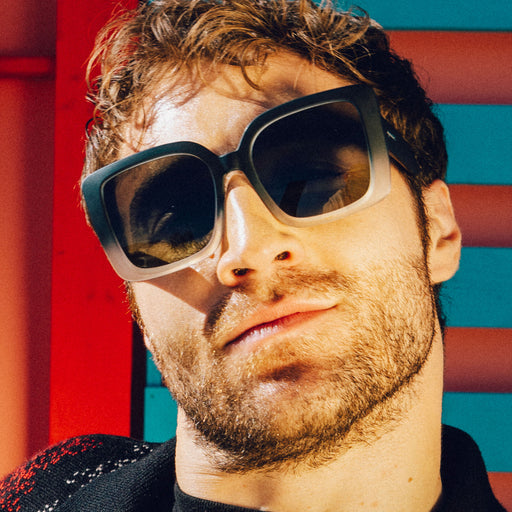 Front view of a young man with brown hair and stubble at a music festival wearing oversized square green shades.