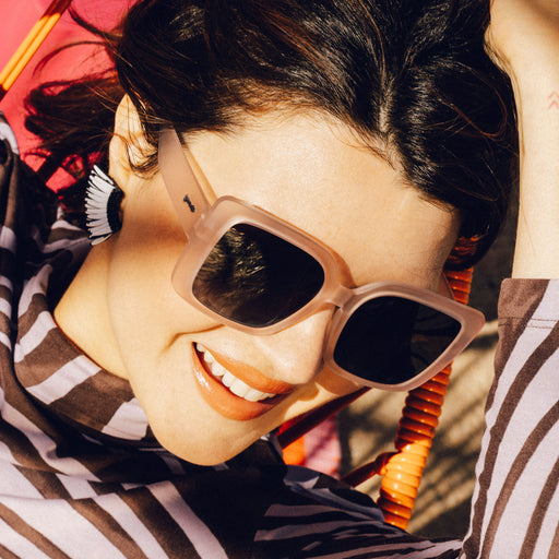 Three-quarter angle view of a young brunette woman wearing oversized square taupe sunglasses laughing at a music festival.