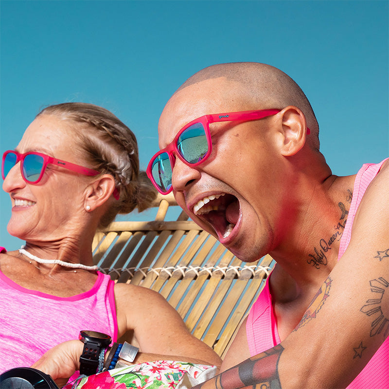 A man and woman wearing hot pink sunglasses with teal lenses laugh while lounging on a tropical beach.