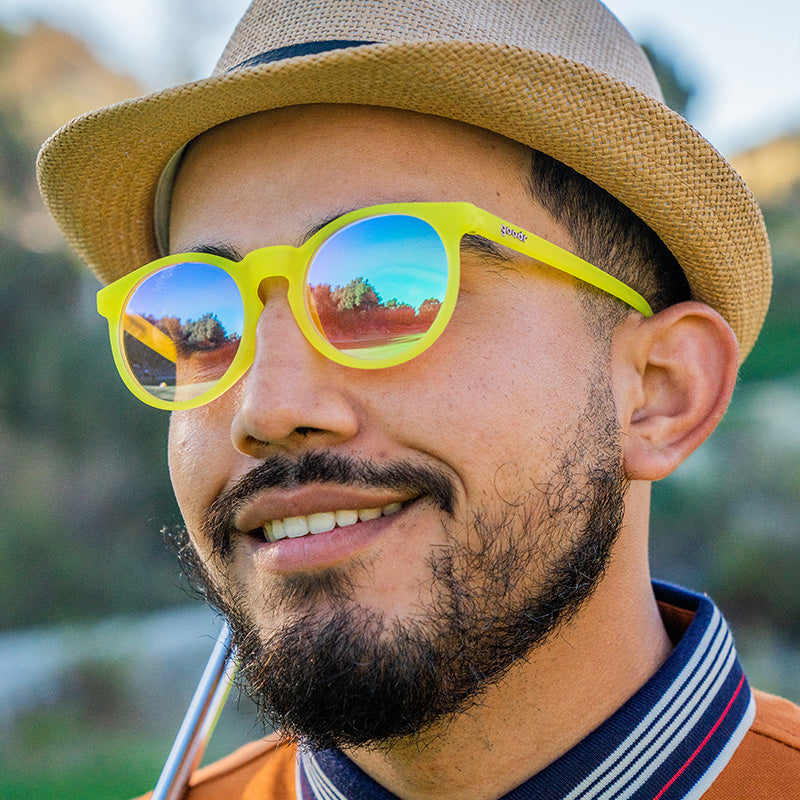A smiling male golfer looks out at the course in round yellow sunglasses with rose-colored circle lenses.