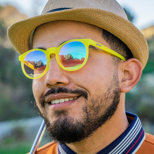 A smiling male golfer looks out at the course in round yellow sunglasses with rose-colored circle lenses.