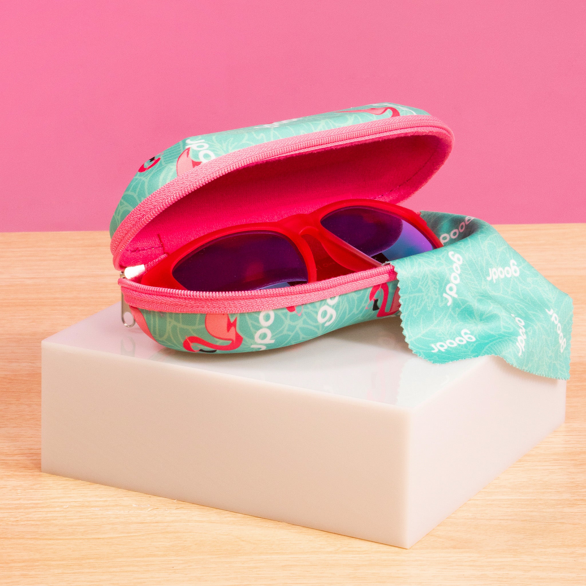 A pair of red-framed sunglasses peeks out of a teal case with pink interior and a zipper, sitting on a light-colored block against a pink background. The case features a playful design with small graphics.