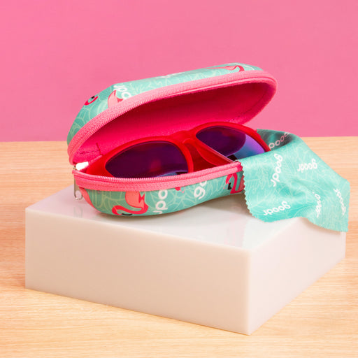 A pair of red-framed sunglasses peeks out of a teal case with pink interior and a zipper, sitting on a light-colored block against a pink background. The case features a playful design with small graphics.