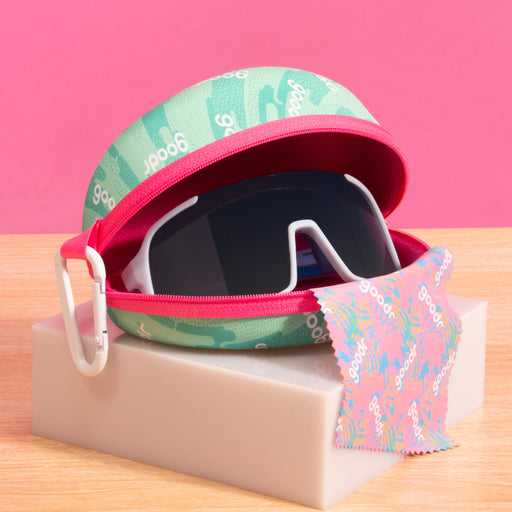 A pair of single lens sunglasses with blue frames and a black lens inside of a pink and teal goodr branded sunglass case, sitting on top of a small white platform on a table.