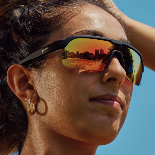 A close-up of a woman wearing reflective wraparound sunglasses with black frames. The lenses have a vibrant orange and yellow mirrored effect, reflecting an outdoor scene with trees and a bright sky.