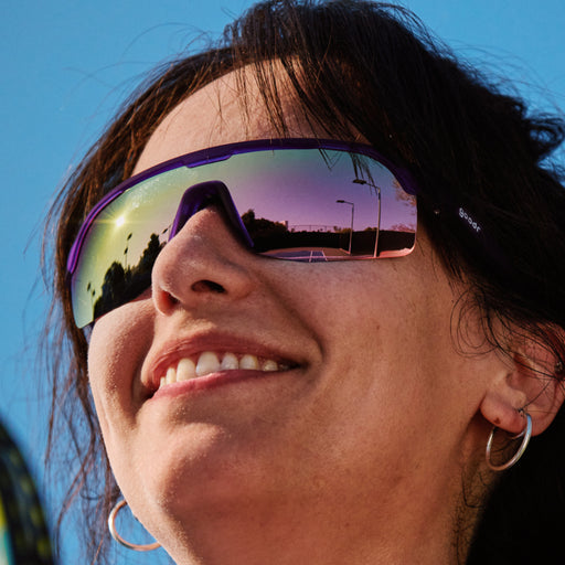 A close-up of a smiling woman wearing large, purple wraparound sunglasses with reflective lenses and a purple frame. The reflection in the lenses shows a sunset scene with streetlights and a road.