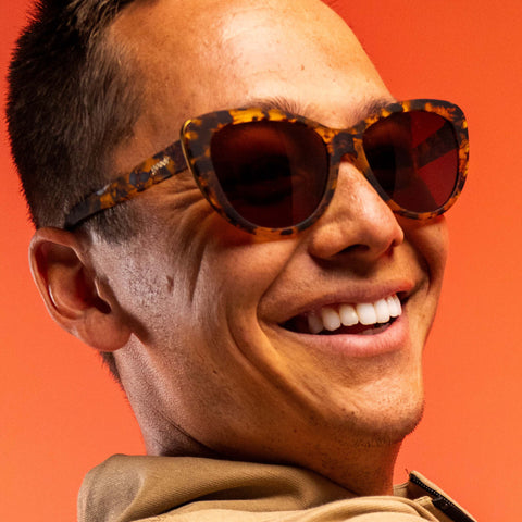 Close-up of a smiling person wearing tortoiseshell sunglasses against an orange background.