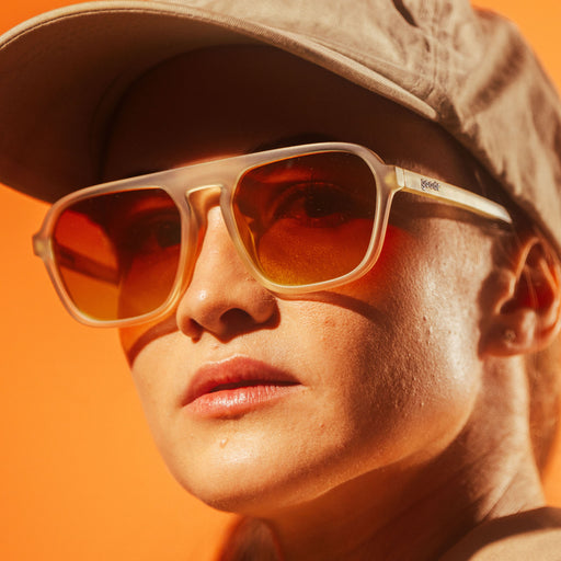 A woman wearing a beige hat, wearing retro aviator, beige sunglasses with reddish-orange, transparent lenses.