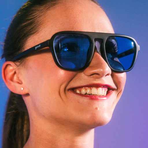 A woman with a pony tail smiling, wearing retro aviator sunglasses with black frames and blue lenses.