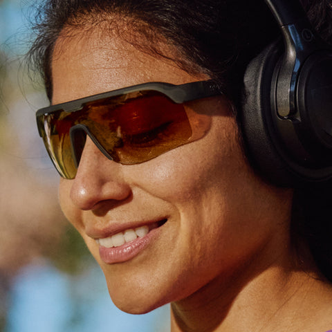 A smiling woman's face, shown wearing black headphones and dark green single-lense sunglasses.