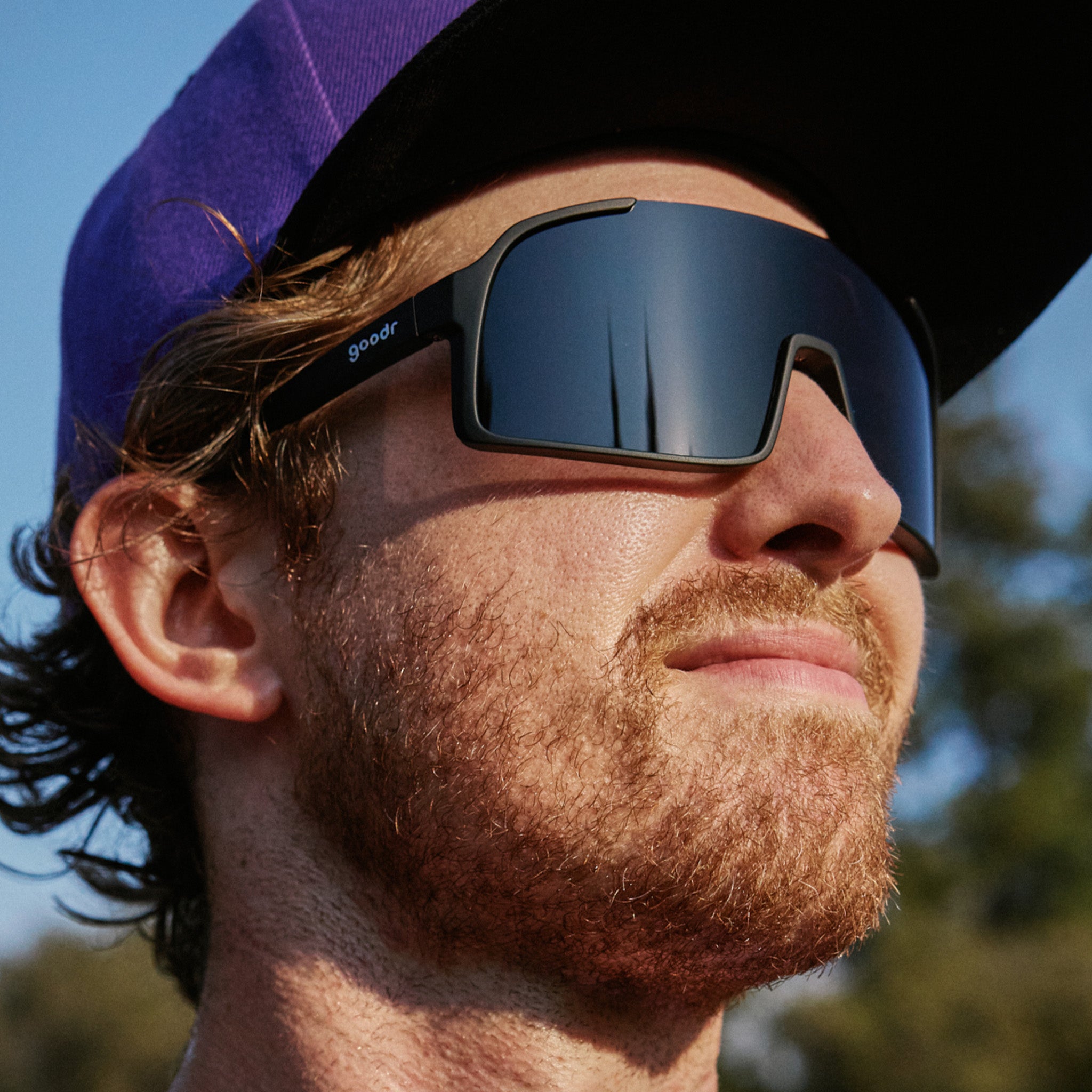 A man standing outside wearing a purple cap and all black, single lens sunglasses.