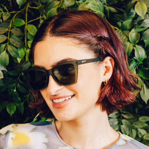 Side profile of a smiling woman wearing translucent dark green angular sunglasses with green non-reflective lenses.