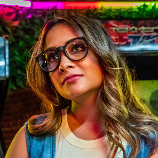 A female gamer wearing round brown tortoiseshell glasses with clear blue-light-blocking lenses looks up at an arcade game.