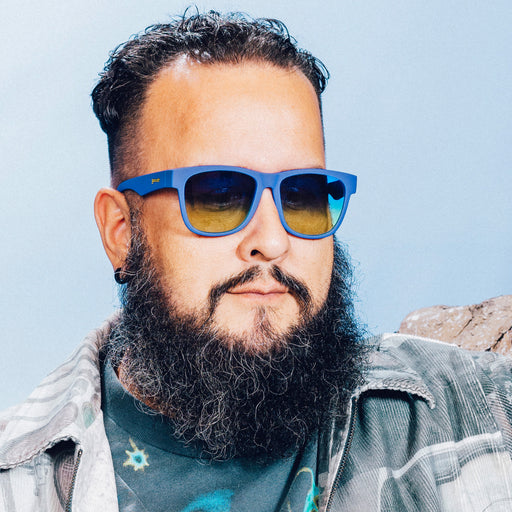 Front view of a man posing in front of a mountain backdrop wearing large blue glasses with blue-yellow ocean gradient lenses.