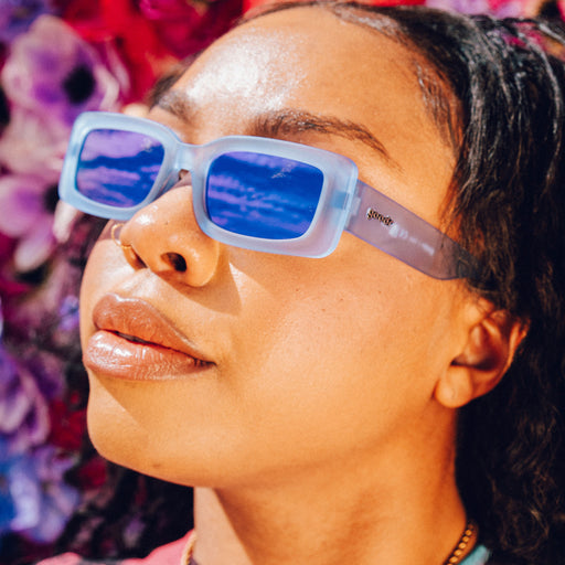 Left profile of a woman wearing and posing with translucent light blue rectangular sunglasses with purple reflective lenses.