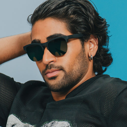 A stylish man wearing sleek matte black half-rim sunglasses with dark green lenses looks off.