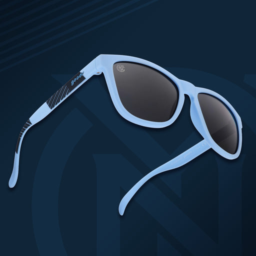 Three-quarter angle front view of NYCFC shades on a blue background. Light blue+navy frames. Black non-reflective lenses.