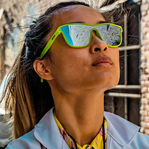 A woman wearing square neon green sunglasses with a single mirrored chrome lens contemplates as she stares off into the distance.
