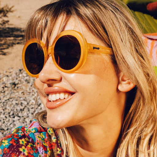Three-quarter profile shot of a woman with medium blonde hair and glossy lips wearing a pair of oversized round orange sunglasses.