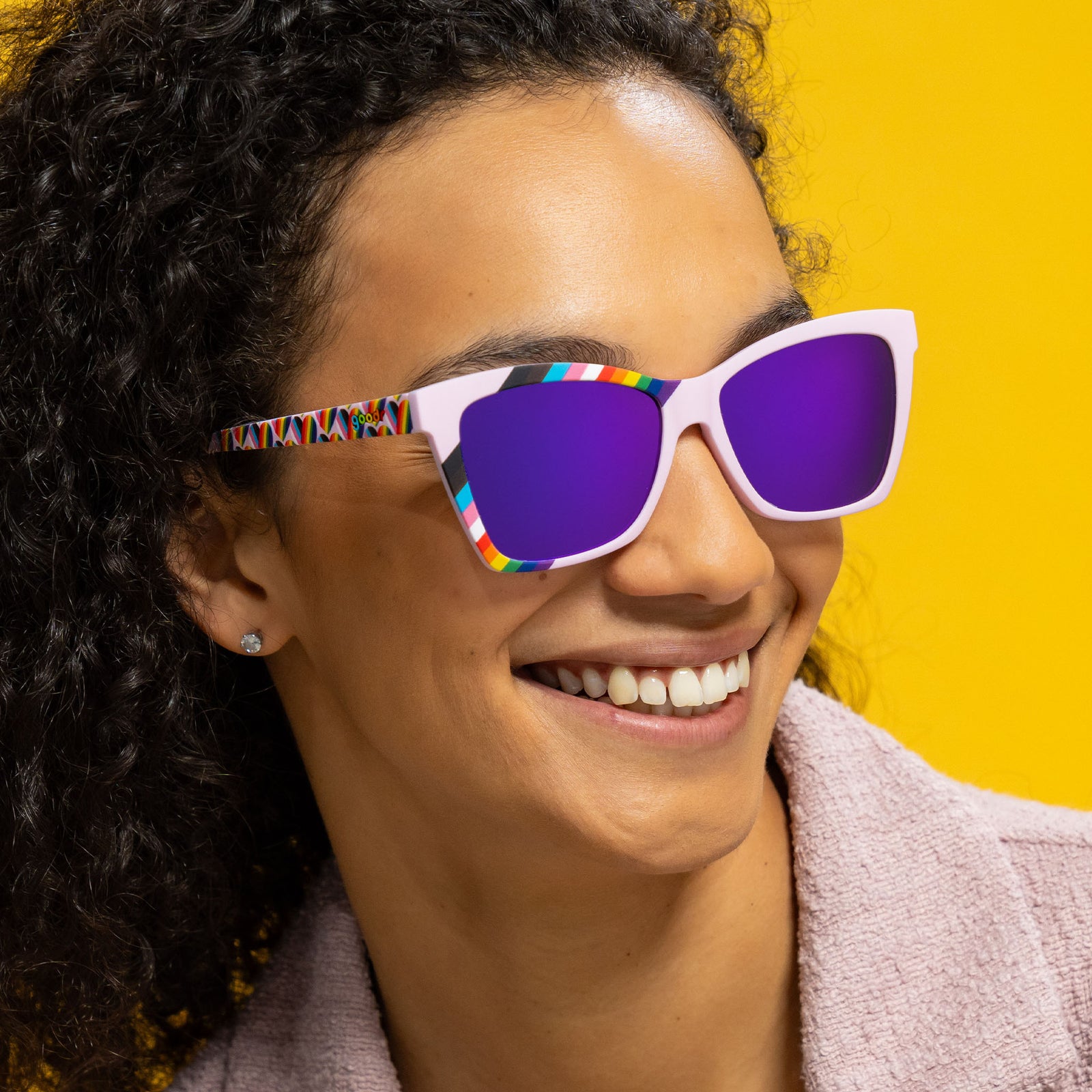 Three-quarter angle view of a happy young woman wearing lilac angled cat-eye LGBTQ Pride sunglasses with purple lenses.