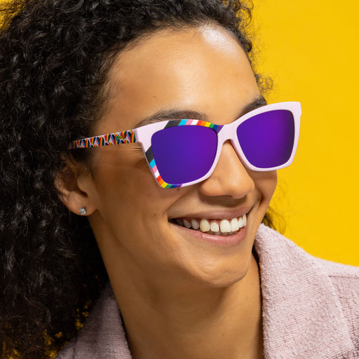 Three-quarter angle view of a happy young woman wearing lilac angled cat-eye LGBTQ Pride sunglasses with purple lenses.