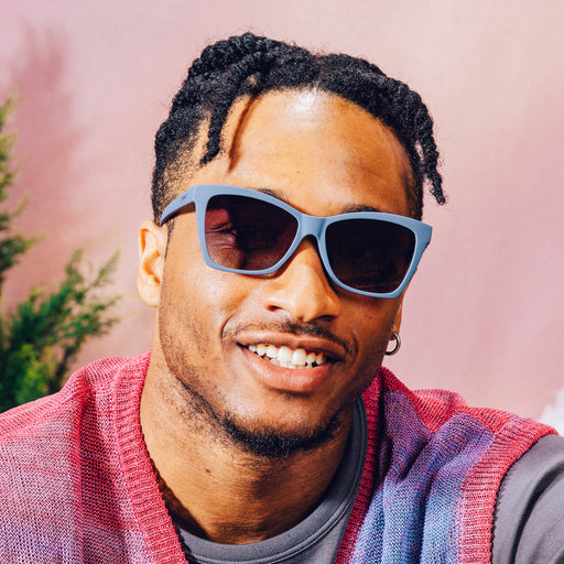 Front view of a man posing wearing dark purple angular sunglasses with purple gradient lenses in front of a meadow backdrop.