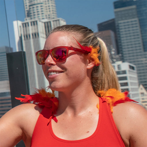 A woman wearing red sunglasses with red lenses with red and orange feather accents, a city skyline behind her.