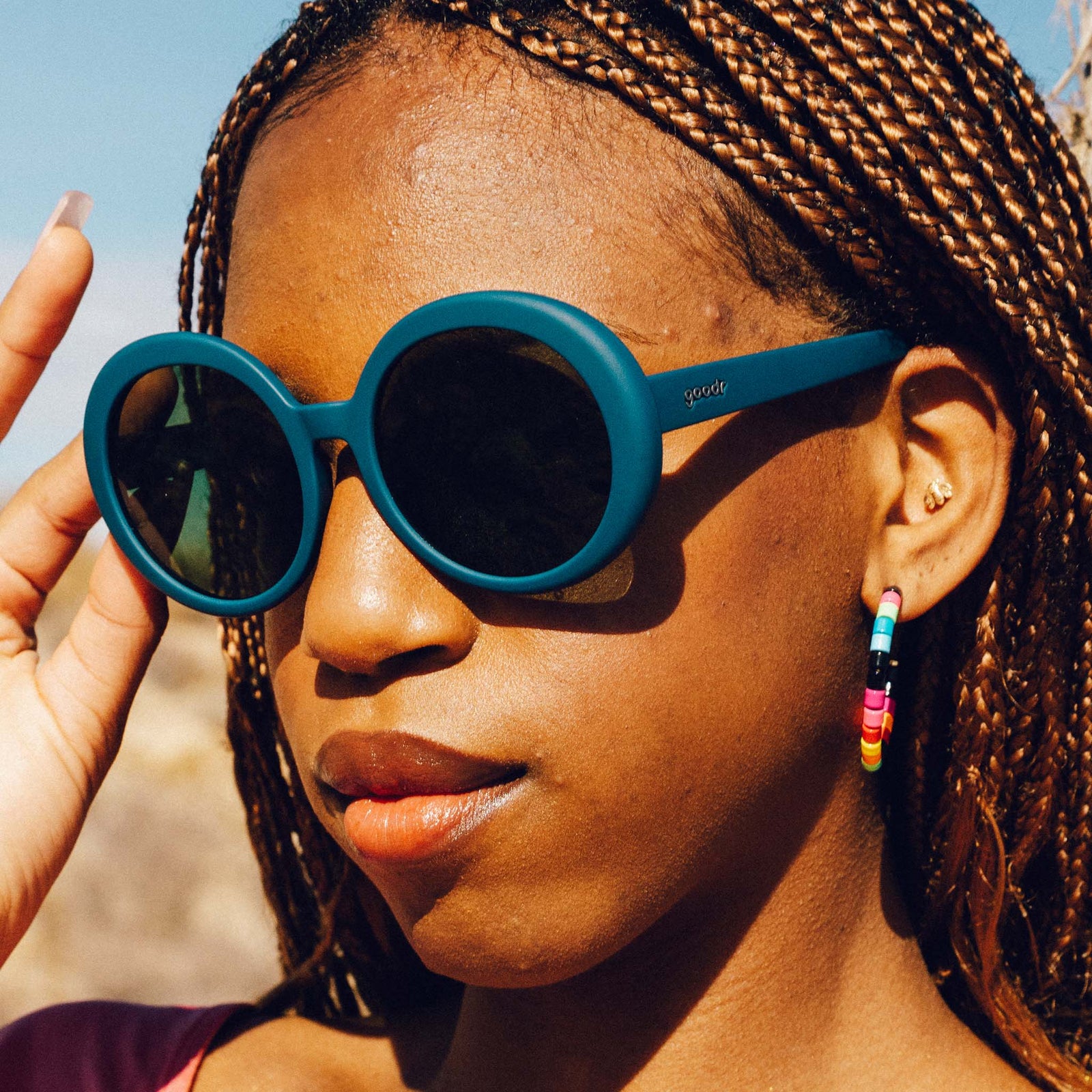 Three-quarter profile of a woman's face. She has copper micro braids, colorful hoop earrings, and oversized round sunglasses in deep green.