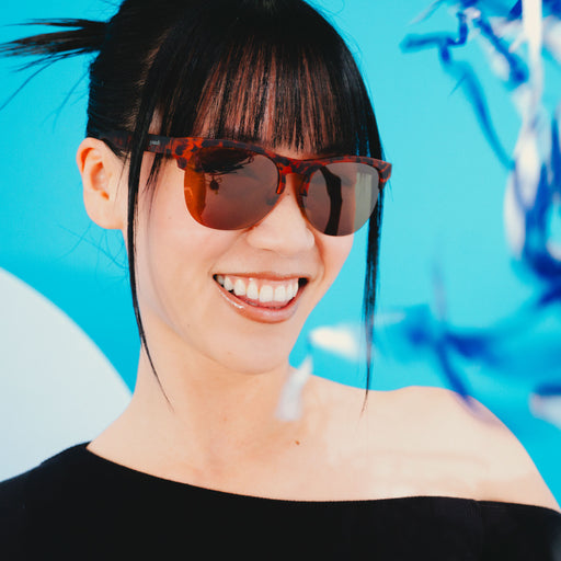 A fashionable woman in sleek brown tortoiseshell half-rim sunglasses with brown non-reflective lenses looks off.