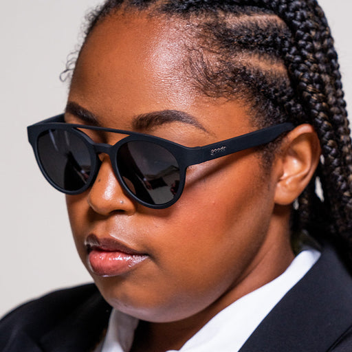 A woman in a sleek black tuxedo and round black sunglasses with a nose bridge and black lenses looks off to the side.