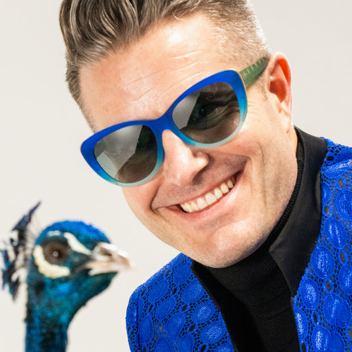A photo of goodr's CEO smiling and wearing a pair of peacock-inspired cat-eye frames. He's posing with a peacock to his left.