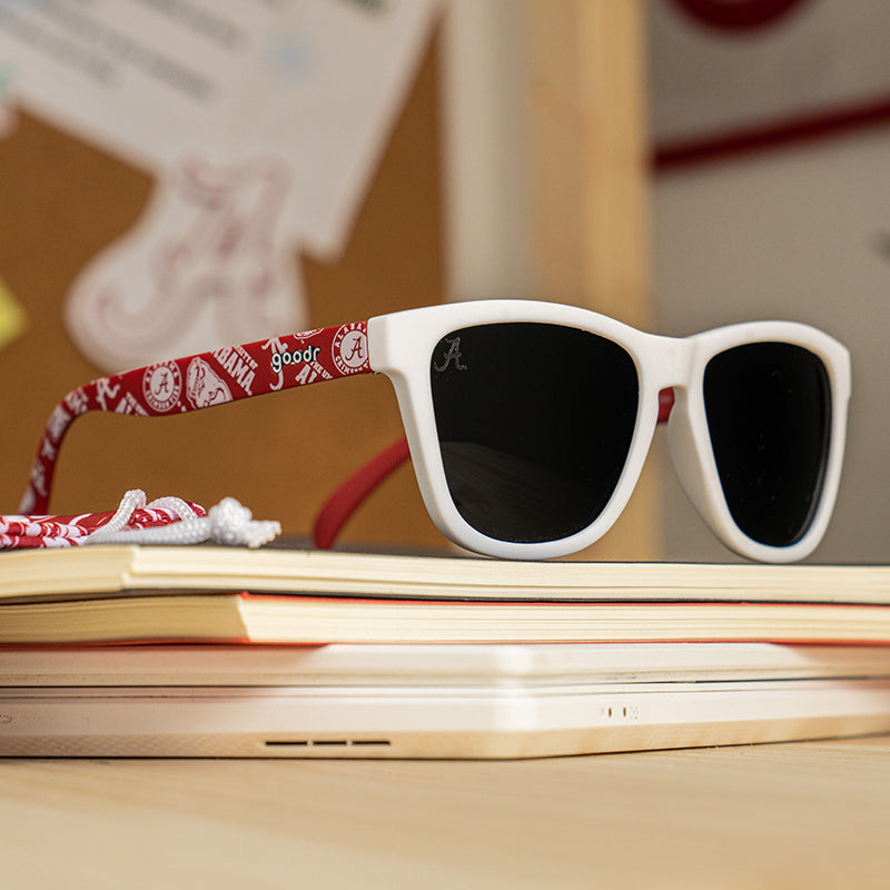 A pair of white-framed sunglasses with dark lenses sits on top of a stack of books. The arms of the sunglasses are decorated with a red University of Alabama-themed pattern, featuring the Alabama "A" logo and other school-related graphics.