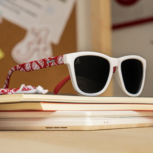 A pair of white-framed sunglasses with dark lenses sits on top of a stack of books. The arms of the sunglasses are decorated with a red University of Alabama-themed pattern, featuring the Alabama "A" logo and other school-related graphics.