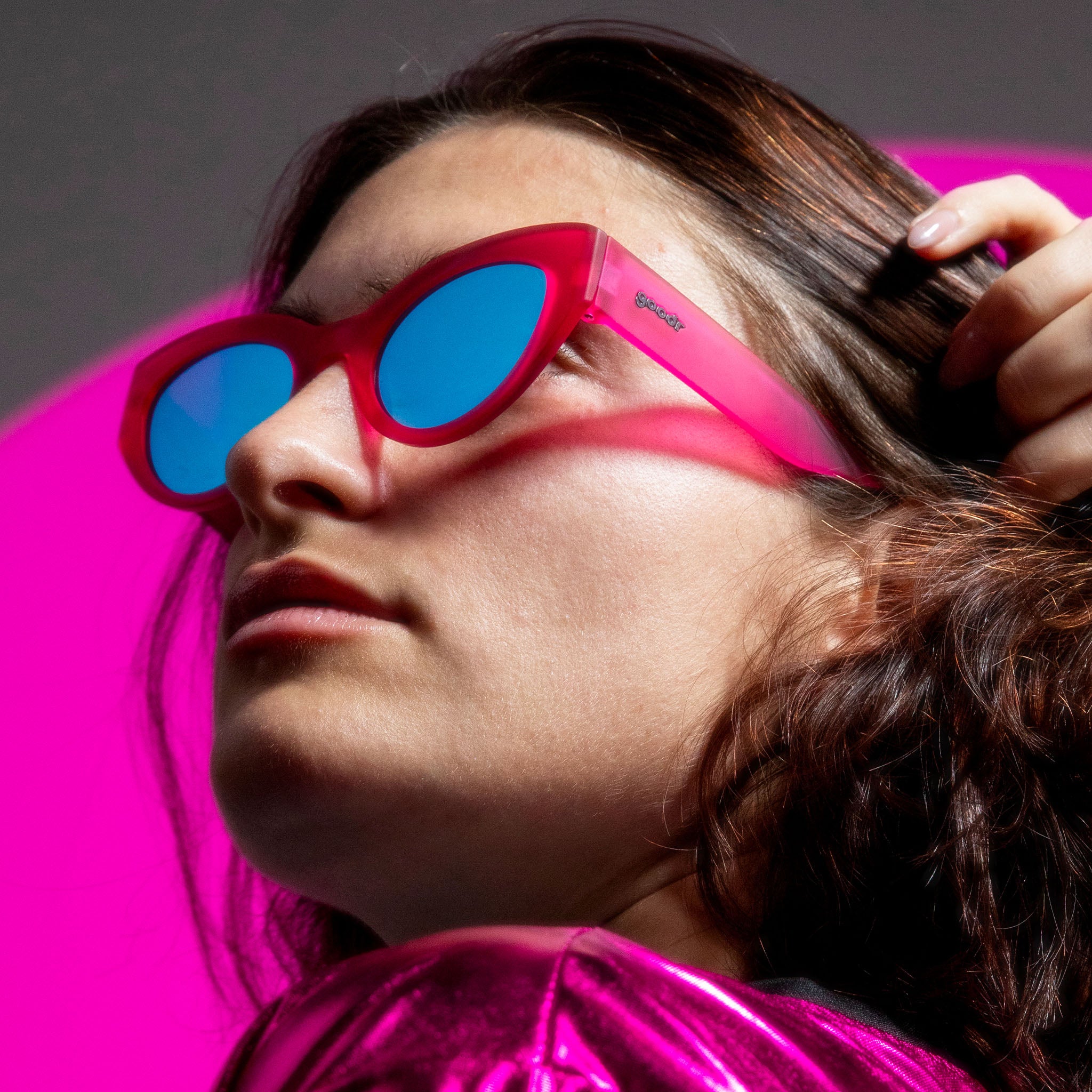 A model with shoulder-length copper hair, tucking it behind their ear and wearing hot pink cat-eye sunnies with blue lenses.