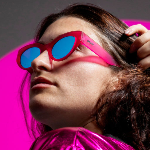 A model with shoulder-length copper hair, tucking it behind their ear and wearing hot pink cat-eye sunnies with blue lenses.