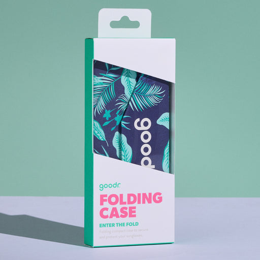 A white product box containing the navy and teal Enter the Fold collapsable sunnies case is displayed on a white tabletop with a sage green backdrop.