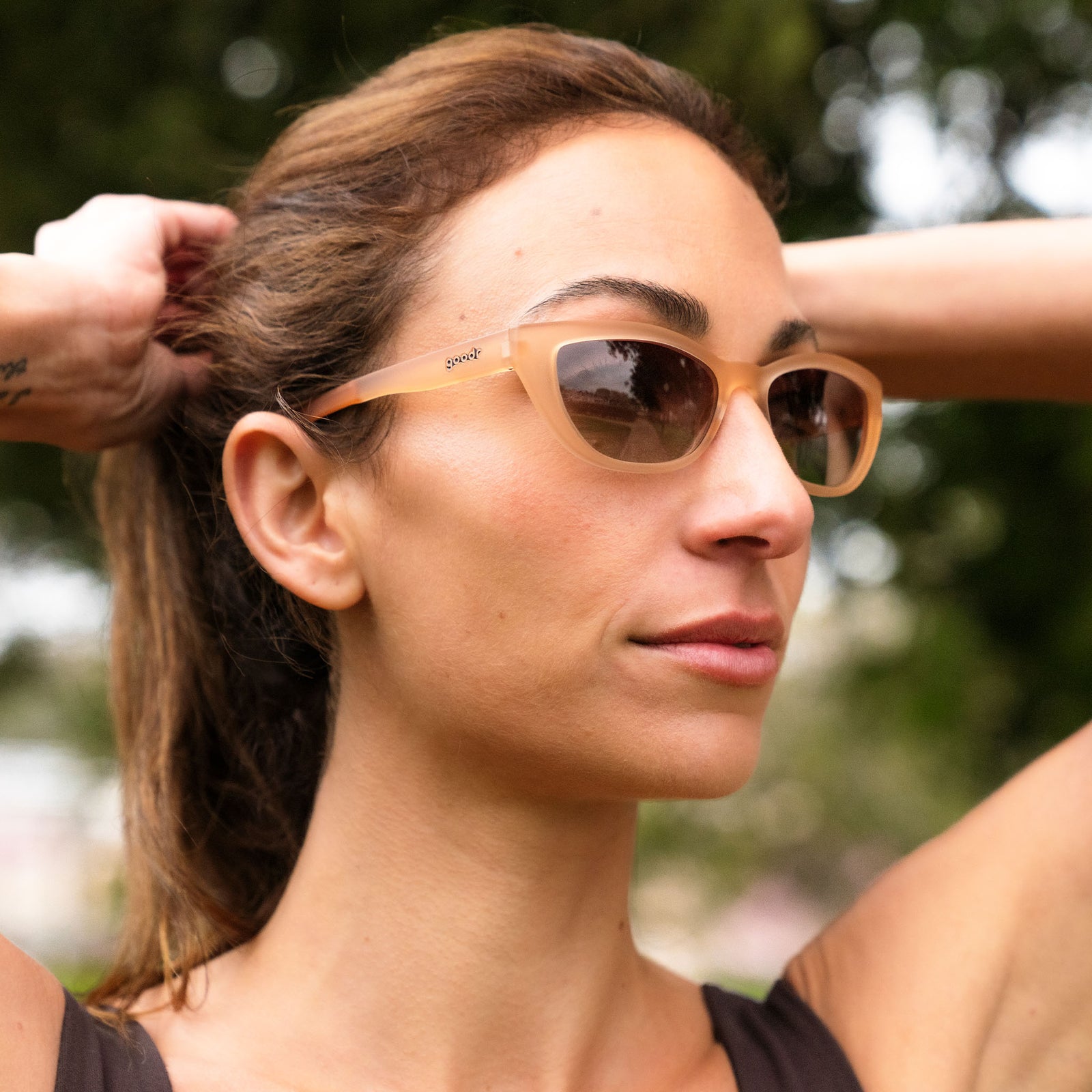 A young woman wearing a brown tank top and translucent brown cat-eye glasses pulls her hair back into a ponytail.