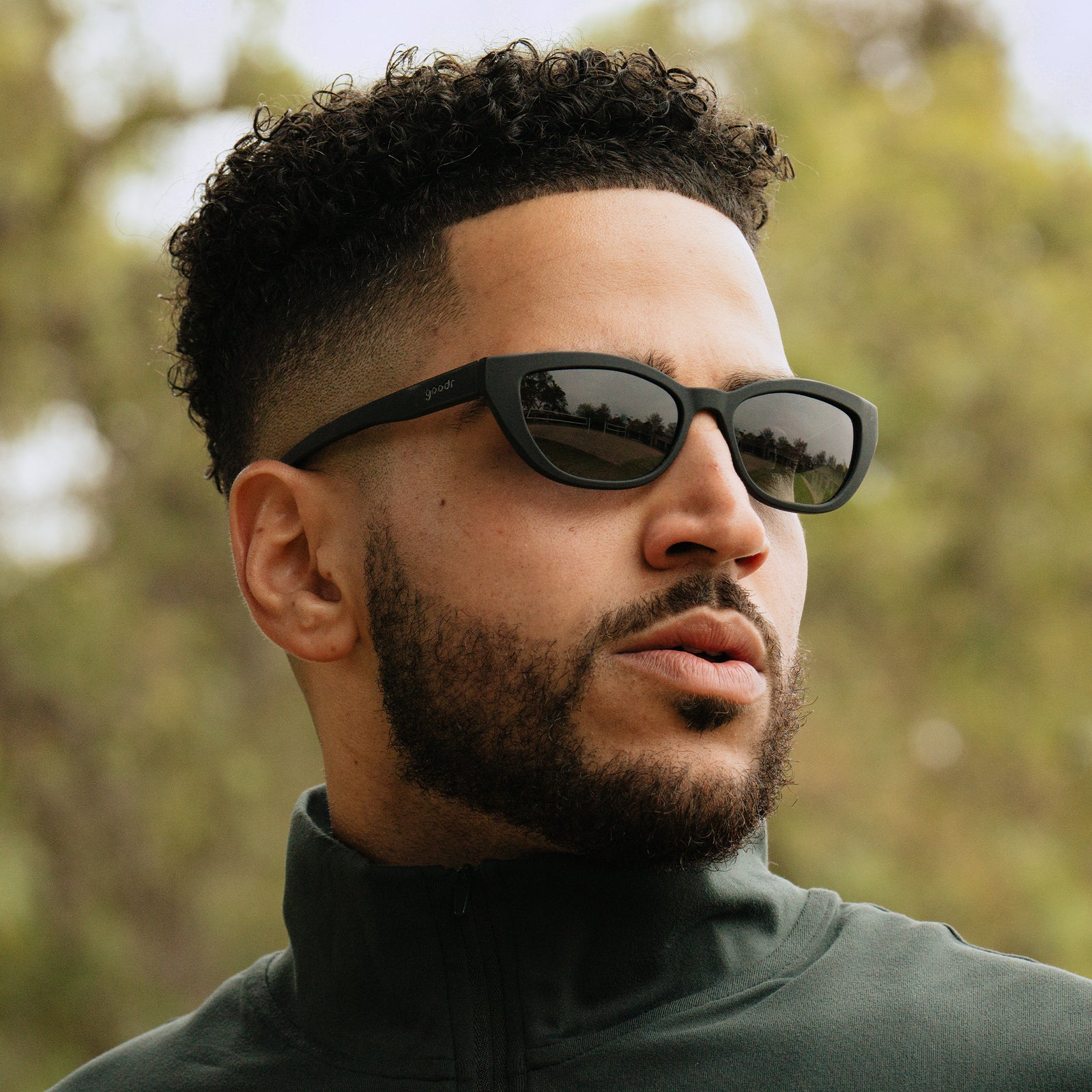 A man with a fade and a dark beard rocks a pair of black cat-eye sunnies, posing against an outdoorsy backdrop of foliage.