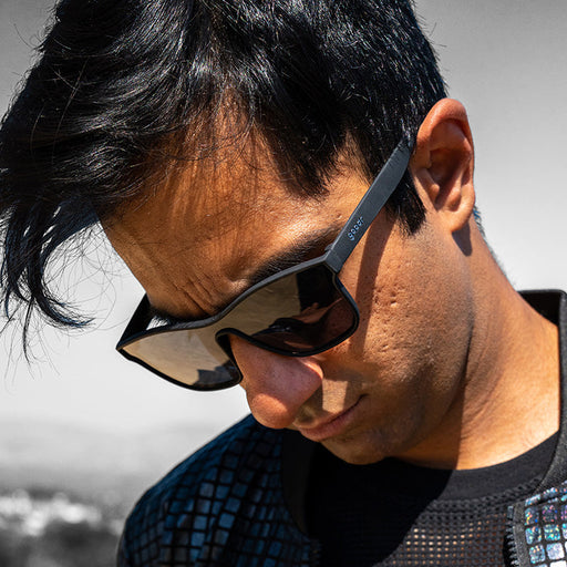 A man wearing black flat single-lens sunglasses looks down to the side, determined.