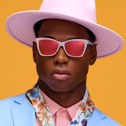 A dapper gentleman poses in a polished outfit of florals, pastels, a fedora, and orange and blue houndstooth Pop G sunnies.