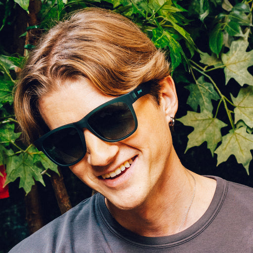Man smiles while wearing large dark teal sunglasses with green gradient lenses in front of a vine backdrop.