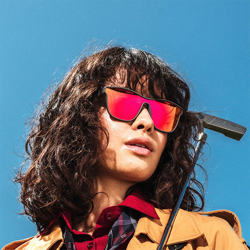 A woman with brown curly hair and bangs, wearing black square shaped sunglasses with reddish-orange reflective lenses