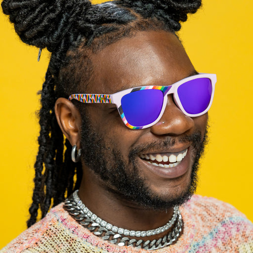 Three-quarter angle view of a happy young man wearing lilac rainbow flag LGBTQ+ Pride sunglasses with purple lenses.