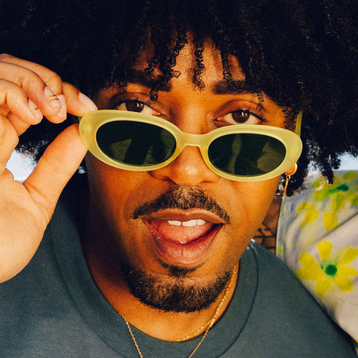 A man with curly black hair and facial hair tilts a pair of citron-colored, oval-shaped sunglasses, perching them on the end of his nose.