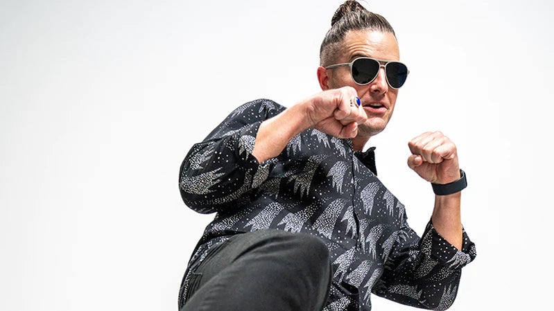 goodr CEO Stephen Lease wearing aviator-style black and grey sunglasses strikes a playful, action-ready pose, with his fists raised and one leg lifted, as if preparing for a kick.